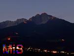29.6.2025, Meran in Sdtirol, Italien. Herz-Jesu-Feuer auf dem Gebirgsgrat des Ifinger Berggipfel.
Der Brauch, im Juni Feuer zu entznden, geht auf frhere Sonnwend- bzw. Johannisfeuer zurck, welche in Erinnerung an das Herz-Jesu-Gelbnis von 1796 umgedeutet wurden. Unten leuchtet das Dorf Tirol in der Dmmerung. 